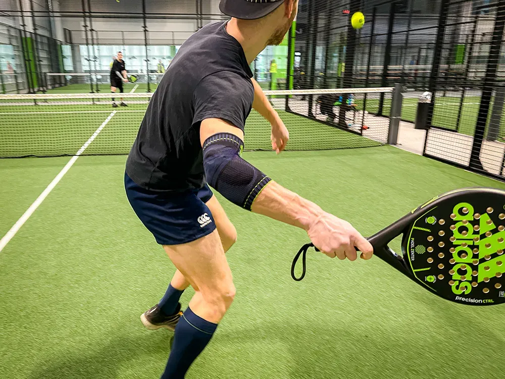 Volledige concentratie tijdens de padelwedstrijd! Een padelspeler in een zwart shirt en korte broek slaat de bal met volle concentratie. Hij draagt een donkerblauwe sportbrace om zijn rechterarm voor extra ondersteuning.