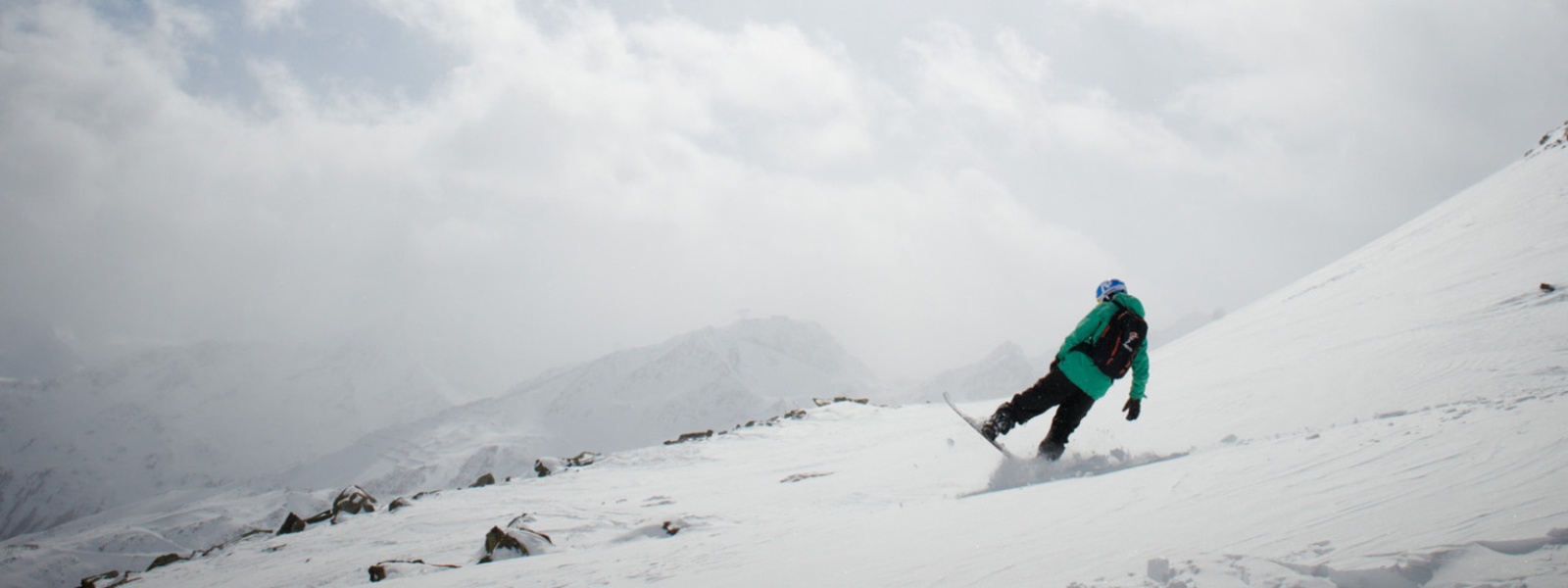 Snowboarders in een groene jasremt met het achterste deel van zijn bord voor een top op de achtergrond, zie je Sneeuwige bergtoppen door de wolken