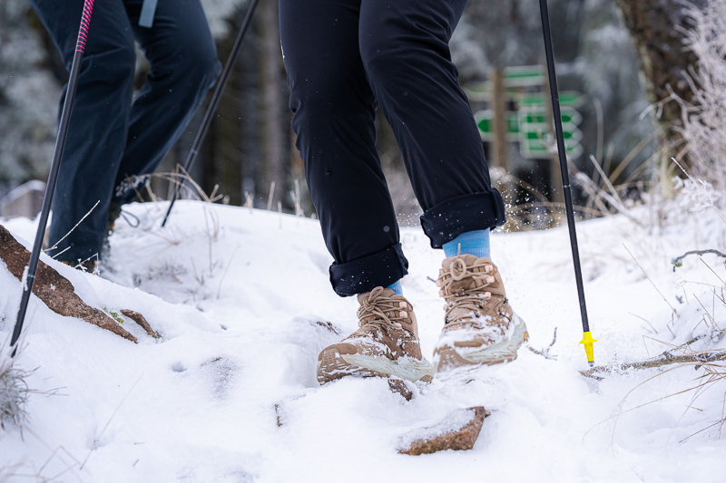 Stap voor stap in de sneeuw – Elke trede telt! Close-up van wandelschoenen die door diepe sneeuw stappen. De voeten behoren toe aan wandelaars met zwarte broeken en blauwe sokken. Wandelstokken met gele punten staan in de sneeuw, terwijl de grond bedekt is met sneeuw en stenen.