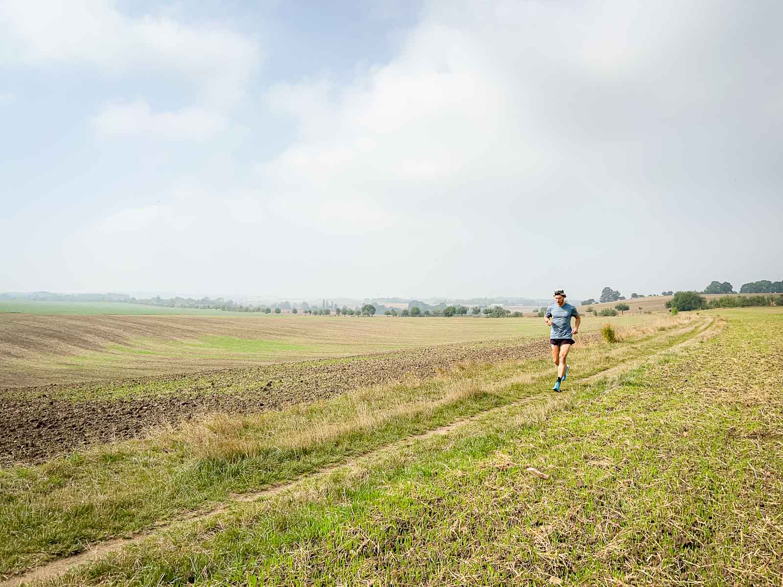 Hardlopen op een Veldweg – Uitgestrekt Landschap voor Duurlopen. Een loper rent op een veldweg in een landelijke omgeving met een lichtbewolkte hemel. De omgeving is rustig en uitgestrekt, perfect voor duurtraining.