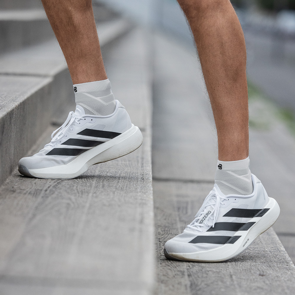 Een man staat met licht gebogen benen op een trap. Zijn witte korte Run Performance Socks en witte hardloopschoenen met zwarte strepen zijn zichtbaar.