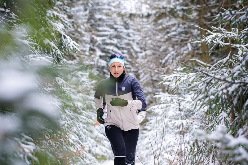 Winterrun: Door besneeuwde sparren op het pad van vastberadenheid. Een hardloopster rent door een besneeuwd bospaadje. De bomen om haar heen zijn zwaar bedekt met sneeuw. Ze draagt een lichtgrijze hardloopjas met zwarte accenten, een zwarte broek en een blauwe hoofdband met een zonnebril.