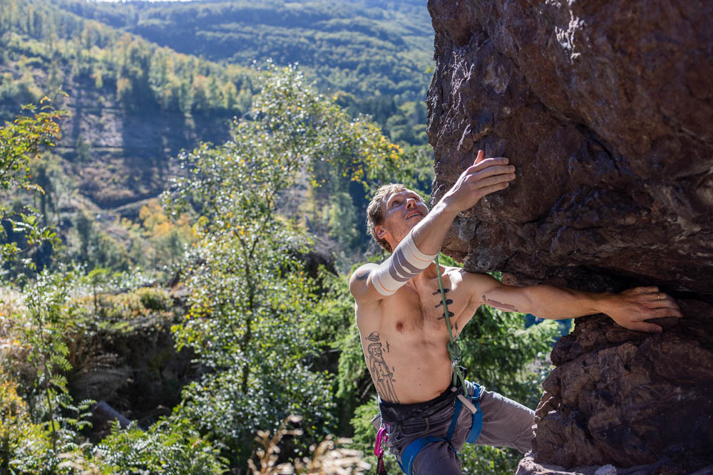 Breed beeld van een klimmer aan een rotsoverhang met uitzicht op het dal; hij draagt de Bauerfeind Sports Outdoor Compression Elbow Support in sandstone terwijl hij zich aan de rots afzet.