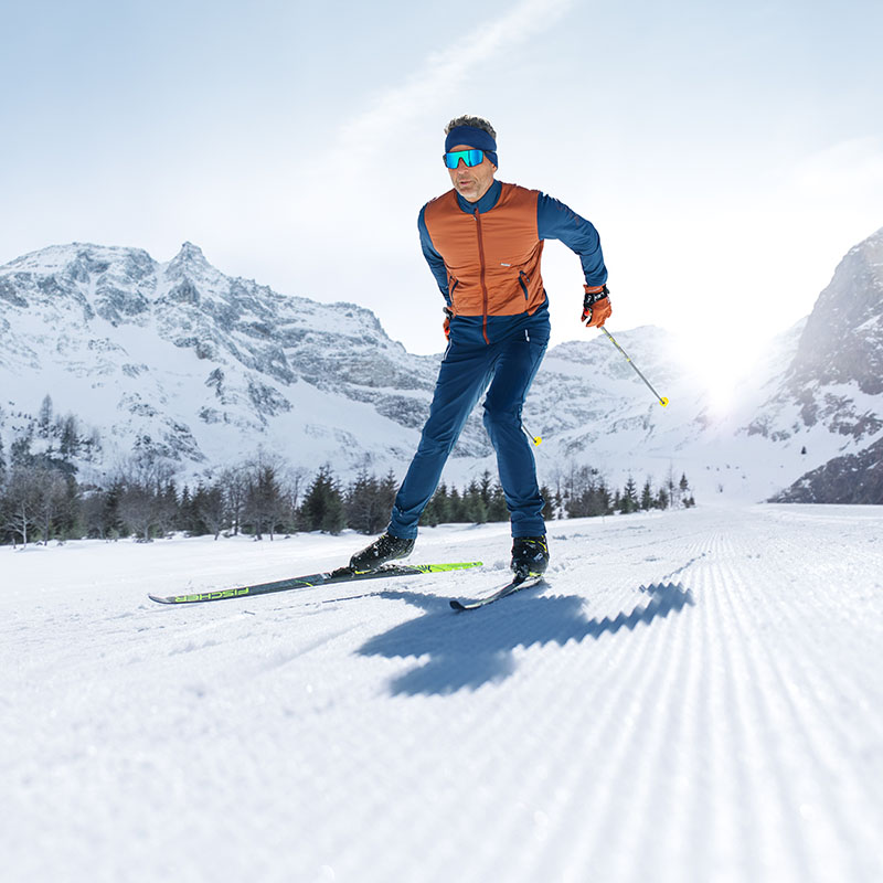 Avontuur op de piste: Skiërs in winterse look Een man met een blauwe skibril, van voren gefotografeerd, is aan het skiën in een winters landschap. Hij draagt een oranje jas en een blauwe skibroek.