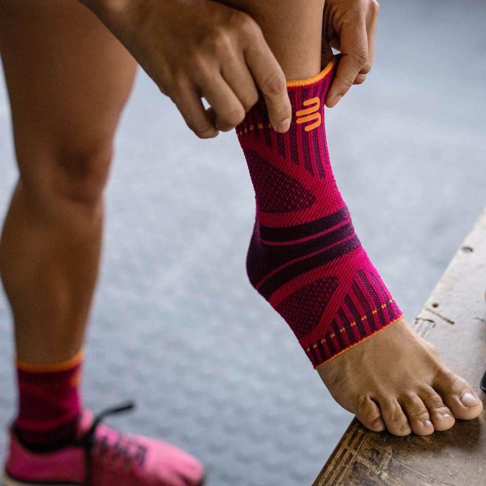 Snel aan, optimale ondersteuning! Close-up van een voet met een roze-zwarte enkelbrace. Een hand trekt de brace over de enkel, terwijl op de achtergrond een roze sportschoen te zien is.