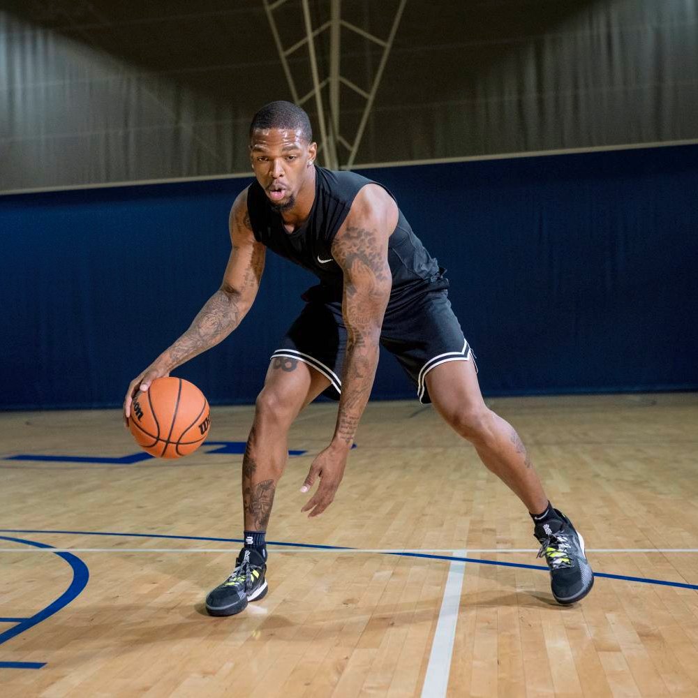 Perfecte controle op het veld – met stevige ondersteuning! Een basketballer in zwarte sportkleding dribbelt op het veld. Hij draagt de Bauerfeind Sports Ankle Support in zwart om zijn enkels.