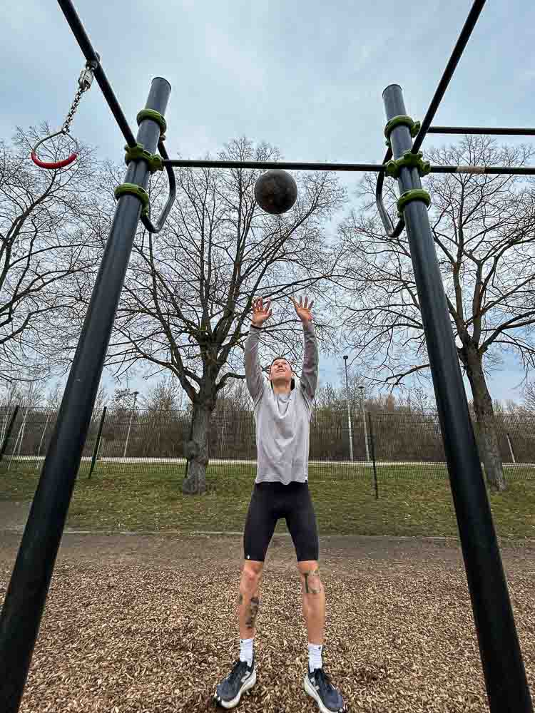 Een sporter gooit een medicine ball omhoog in een outdoor trainingspark onder een calisthenics-rek. De scène laat een functionele kracht- en conditieoefening zien die goed past bij HYROX-training zonder sportschool.