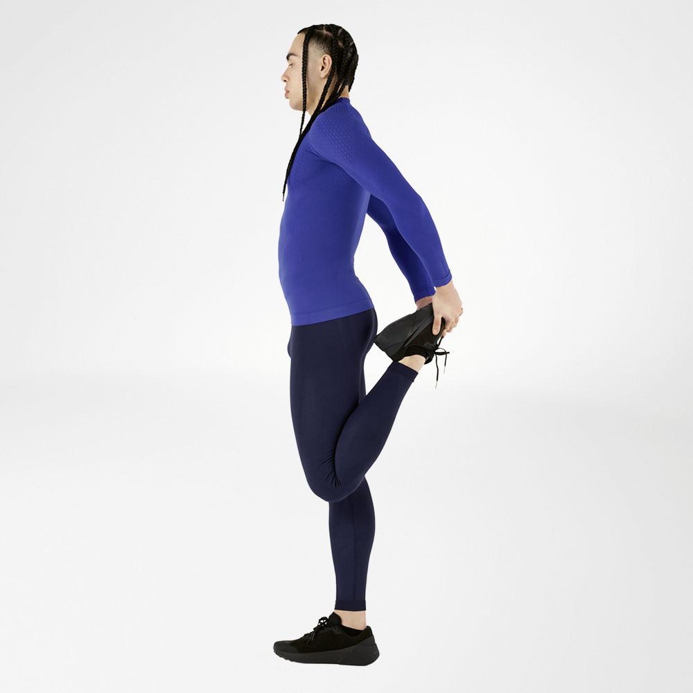 Een atleet in een blauw compressie-sportoutfit, bestaande uit een shirt met lange mouwen en tights, voert een rekoefening uit en houdt één been naar achteren, tegen een witte achtergrond. Het outfit biedt hoog comfort en optimale spierondersteuning.