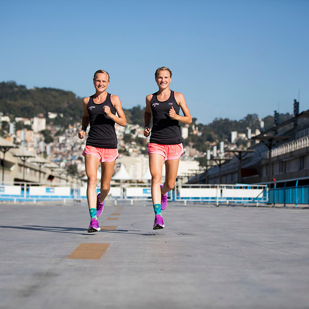 Hahner Twins rennen op een straat met een glimlach op de camera om enkelbandages voor sport te dragen. Een stad en heuvel zijn op de achtergrond te zien.