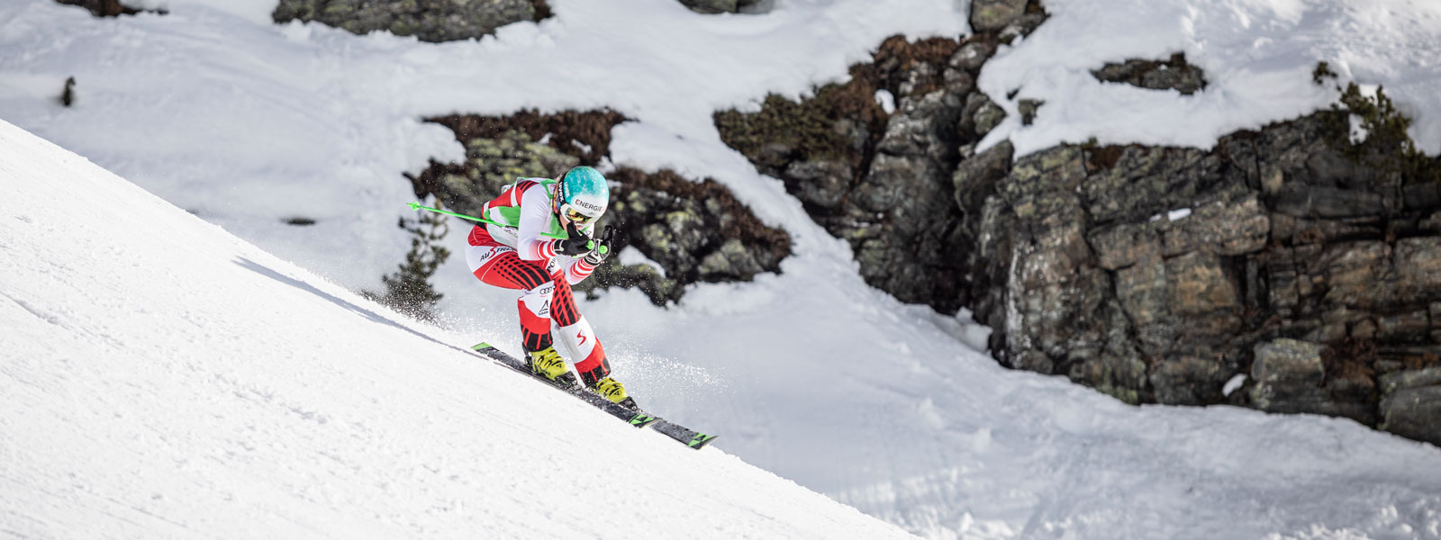 Ski-hardloper in een rood en wit racepak in een bergafwaarts positie met een lichte schommel met grote met sneeuw bedekte rotsen op de achtergrond