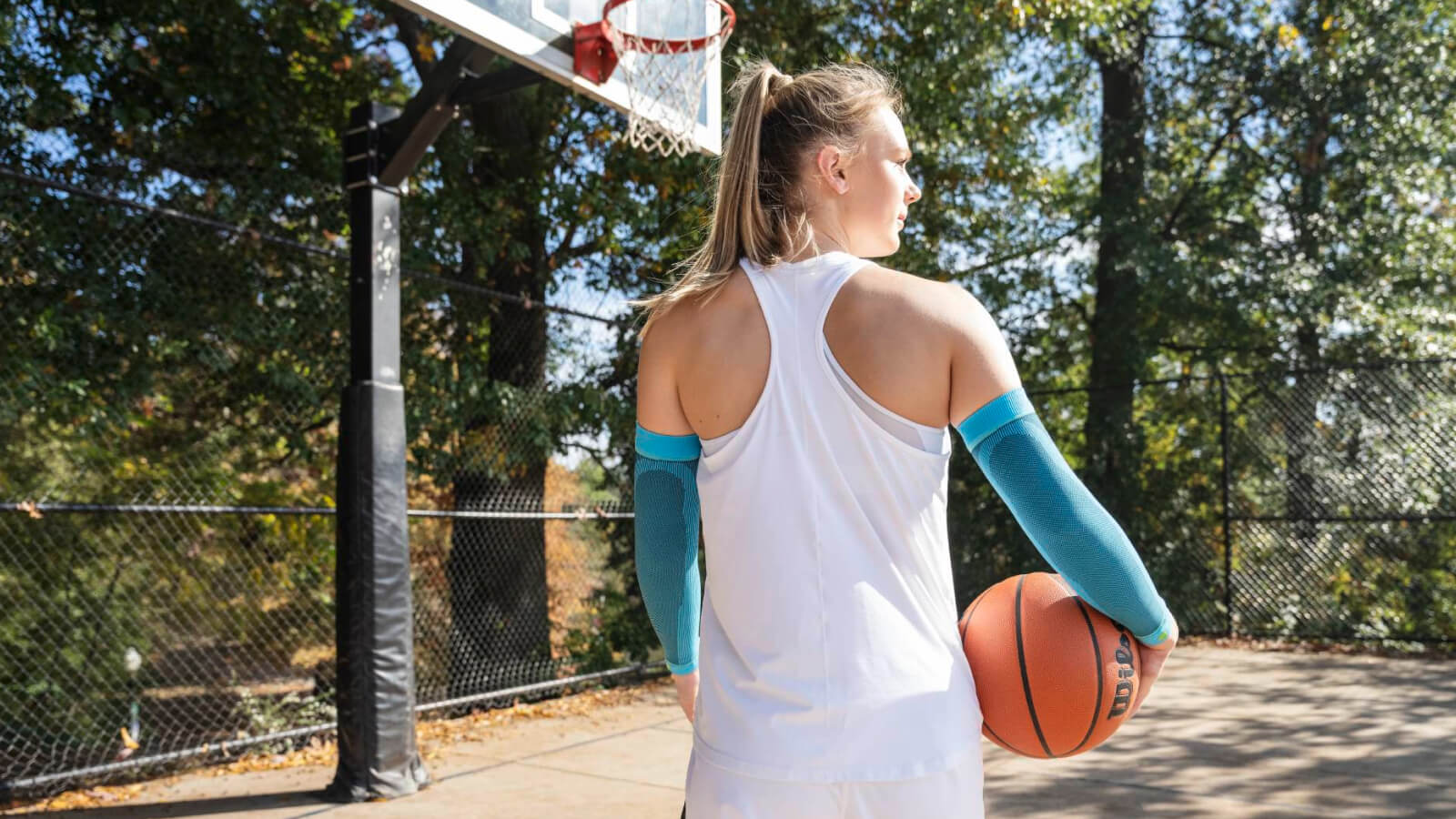 Klaar voor de volgende worp. Een vrouwelijke basketbalspeelster staat op een buitenveld met een basketbal in haar rechterhand. Ze draagt lichtblauwe compressie-armhoezen en kijkt geconcentreerd naar de basket.