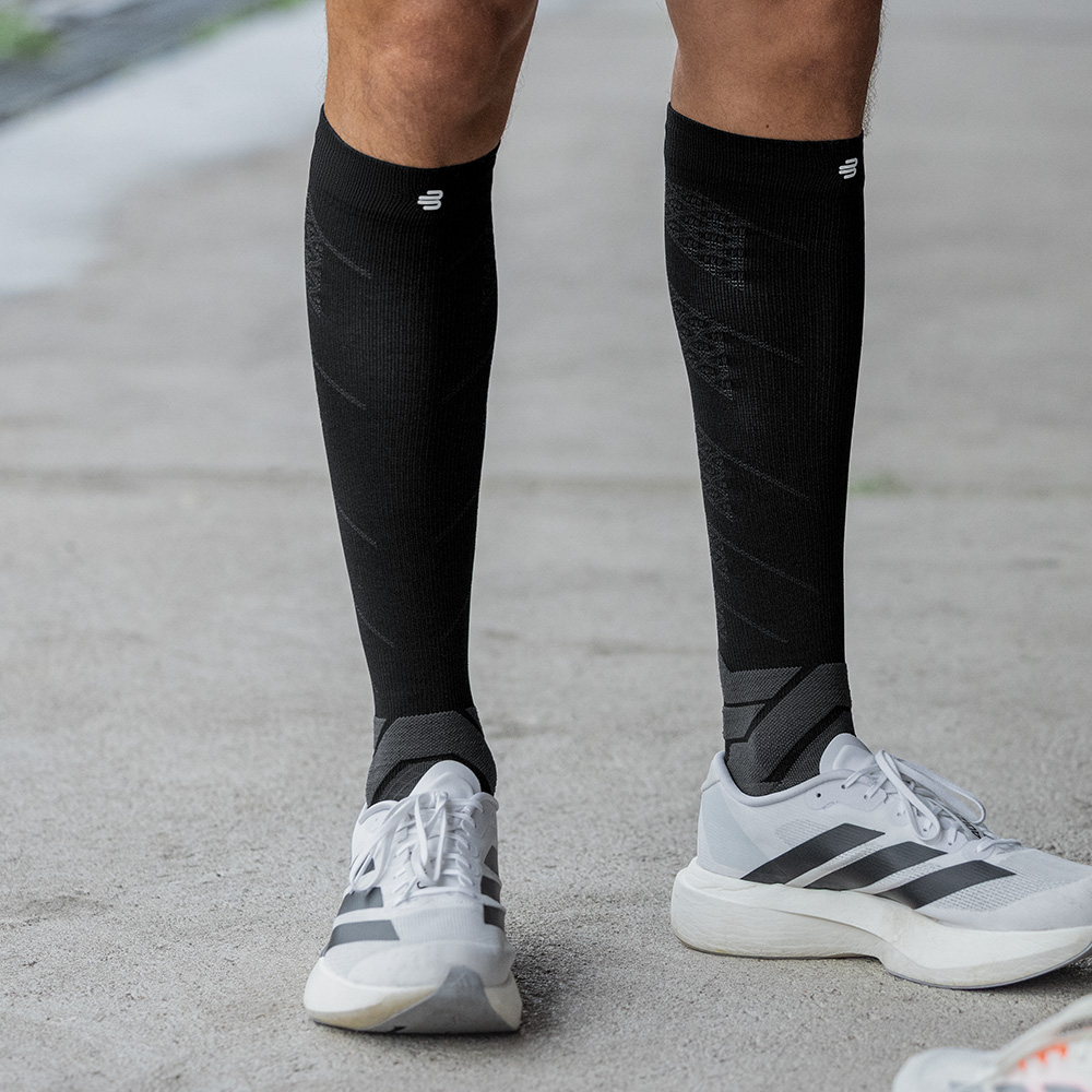 Een sportieve man staat rechtop op asfalt, met zwarte compressiesokken van Bauerfeind en witte hardloopschoenen. Zijn rechte houding en gespannen kuitspieren benadrukken het stabiliserende effect van de sokken tijdens het staan.