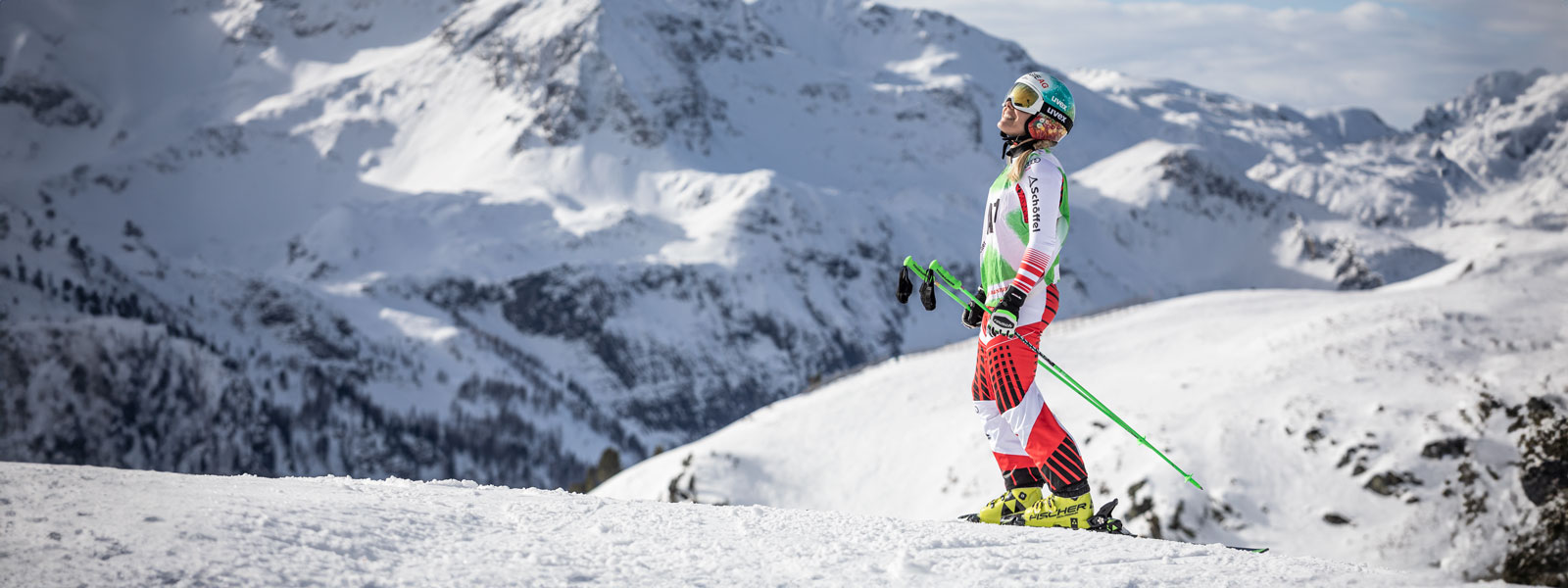 Ski -chauffeur in een rood en wit racepak glimlacht en met de stokjes in haar handen voor een enorme bergketen