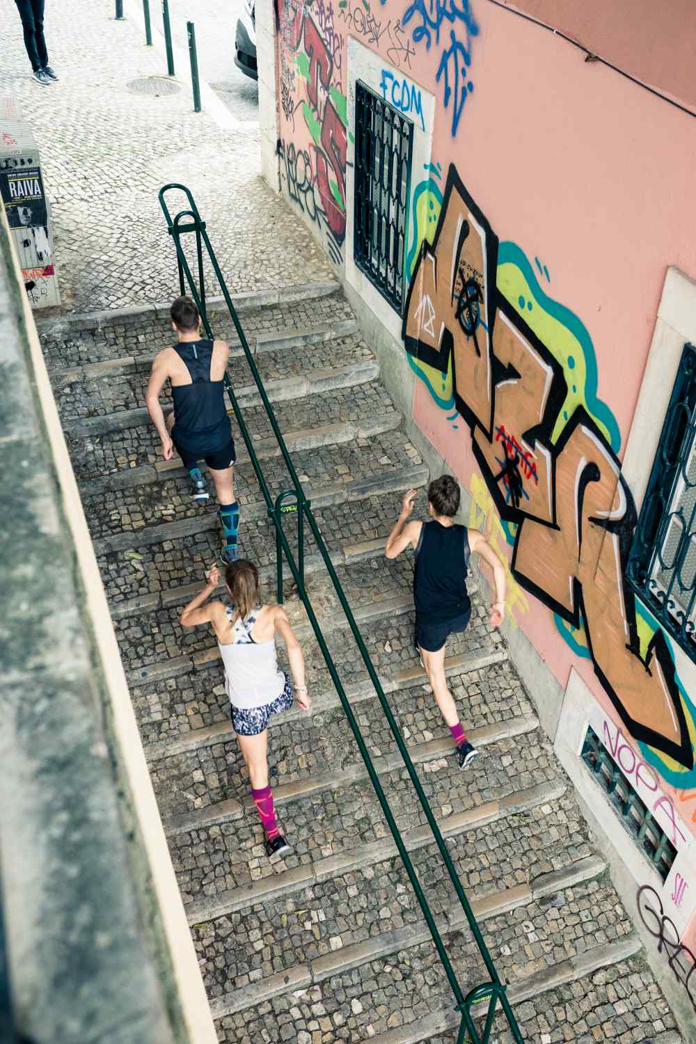 Bird's Eye View: een man en twee vrouwen in rennende outfits lopen een trap in een oude stad met graffiti op de muren