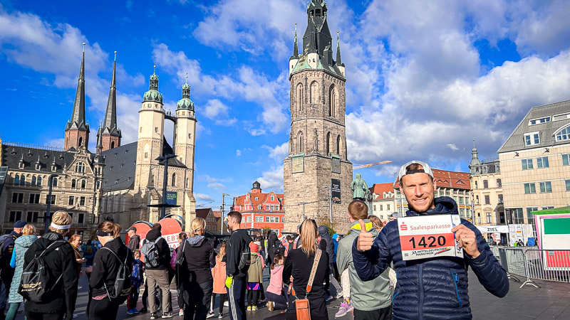 Deelnemer bij het startpunt van de Mitteldeutscher Halbmarathon op de marktplaats van Halle (Saale) met de Marktkirche en de Rode Toren op de achtergrond. Een deelnemer van de Mitteldeutscher Halbmarathon houdt zijn startnummer omhoog op de marktplaats van Halle (Saale). Achter hem zijn de historische Rode Toren en Marktkirche te zien. Op de voorgrond bereiden lopers en toeschouwers zich voor op de race.