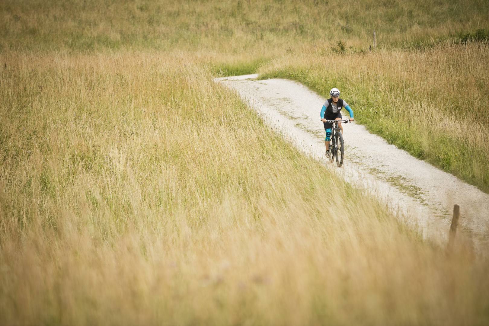 Vrouw met armsleeves en kniebrace op de achtergrond van het beeld loopt op een mountainbike over een pad tussen hoog gras