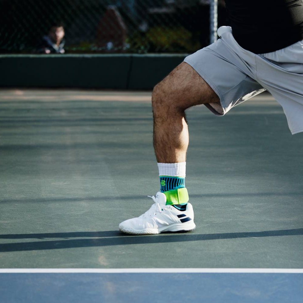 Een tennisspeler op de hardcourt, in beweging tijdens een opslag. Hij draagt de Bauerfeind Sports Ankle Support Rivera in blauw-groen over zijn witte sportsokken.