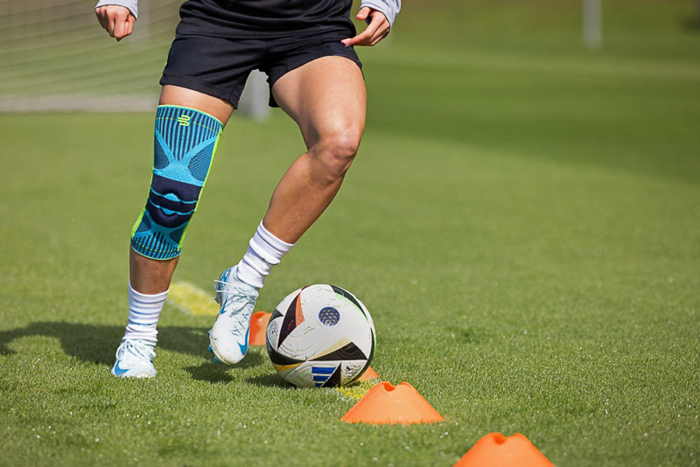 Primo piano di una calciatrice che indossa calzini a compressione Bauerfeind, con un piede appoggiato su un pallone. Il prato verde e i dettagli nitidi mettono in risalto le prestazioni atletiche e la qualità dell'attrezzatura.