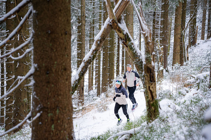 Winterse looptocht onder de boog van de natuur Twee hardlopers rennen door een besneeuwd bos. Het pad wordt omringd door hoge met sneeuw bedekte sparren, terwijl een natuurlijke boog van takken over het pad buigt. Beiden dragen warme hardloopkleding en hoofdbanden.