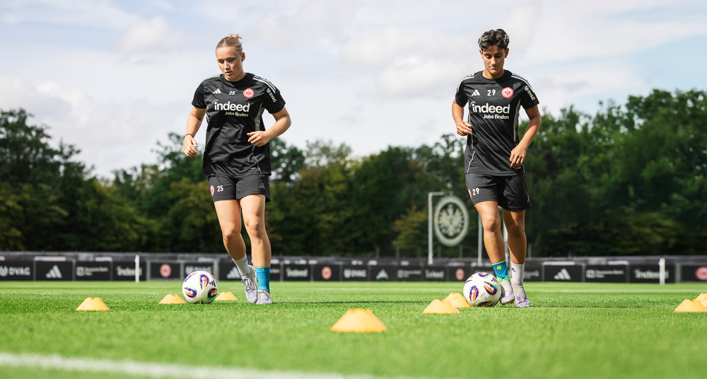Twee speelsters van Eintracht Frankfurt doen een coördinatietraining met pionnen en bal, gekleed in zwarte trainingsoutfits.