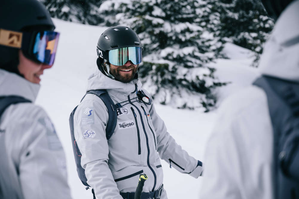 Goede, nou ja -being skiër met een zwarte helmgrijze jas en backpack -opname tussen twee andere skiërs