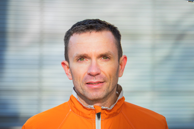 Een portret in de zon: Christian Seiler Een man in een feloranje jas glimlacht naar de camera. Op de achtergrond staat een modern gebouw met bomen en bloeiende planten.
