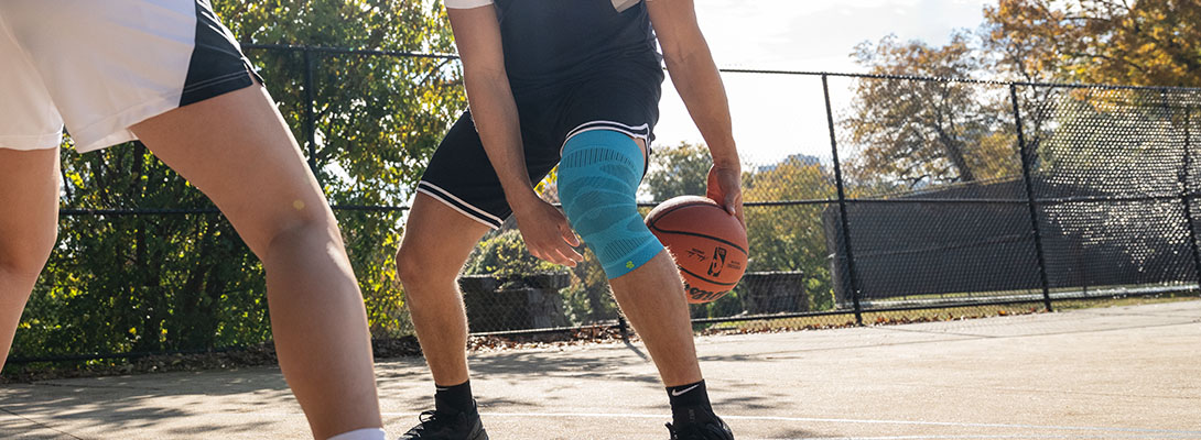 Kniehoes bij basketbal Een basketbalspeler staat op een betonnen veld dat is omgeven door een hek en doet een truc met de bal door hem door zijn twee benen te laten gaan. Een andere speler staat tegenover hem. Hij draagt een kniekous.