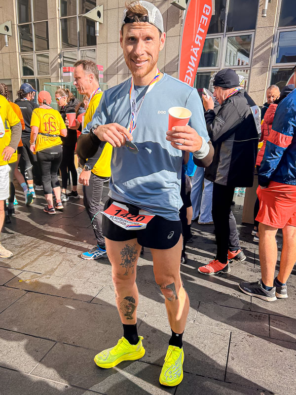 Bij de finish van de halve marathon van Halle, trots met medaille en drankje Trots bij de finish na het voltooien van de halve marathon in Halle, met een medaille en een drankje in de hand, genietend van het moment na de race.
