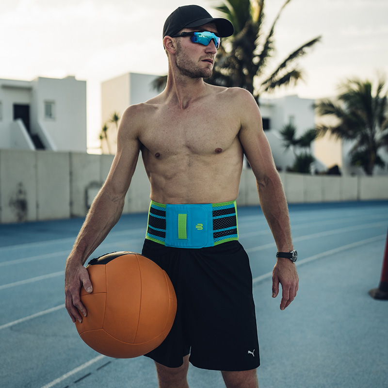 Sterke rug, sterke training – haal het beste uit jezelf met stabiele ondersteuning. Een sportieve man met een pet en zonnebril staat op een atletiekbaan en draagt een blauwe rugbrace met groene accenten. Hij houdt een grote medicijnbal in zijn hand.