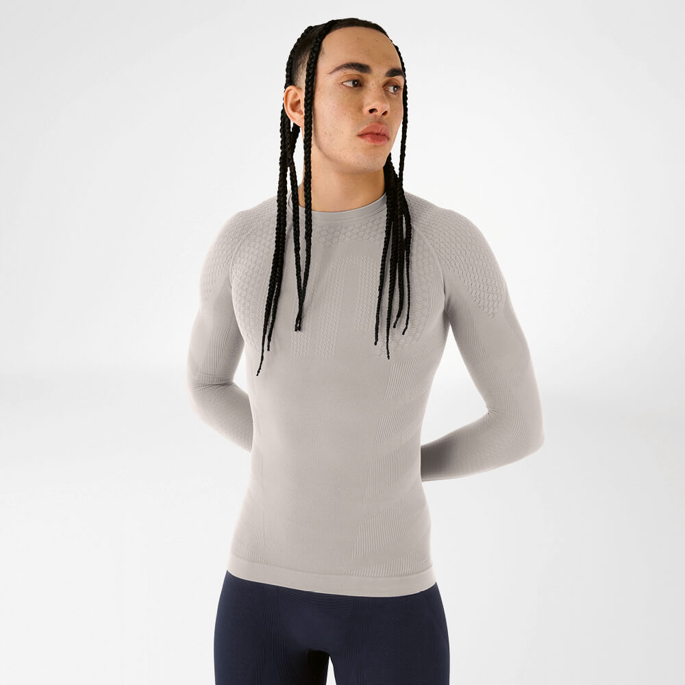 Klaar om te presteren – Compressieshirt in topvorm en stijl Model met lange vlechten draagt een beige compressieshirt met lange mouwen voor een neutrale achtergrond. De structuur benadrukt het naadloze, functionele design.