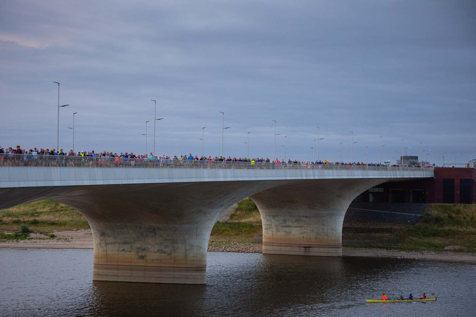 Honderden deelnemers van de 4Daagse lopen bij zonsopgang over een brede brug in Nijmegen, terwijl onder hen een roeiboot passeert.
