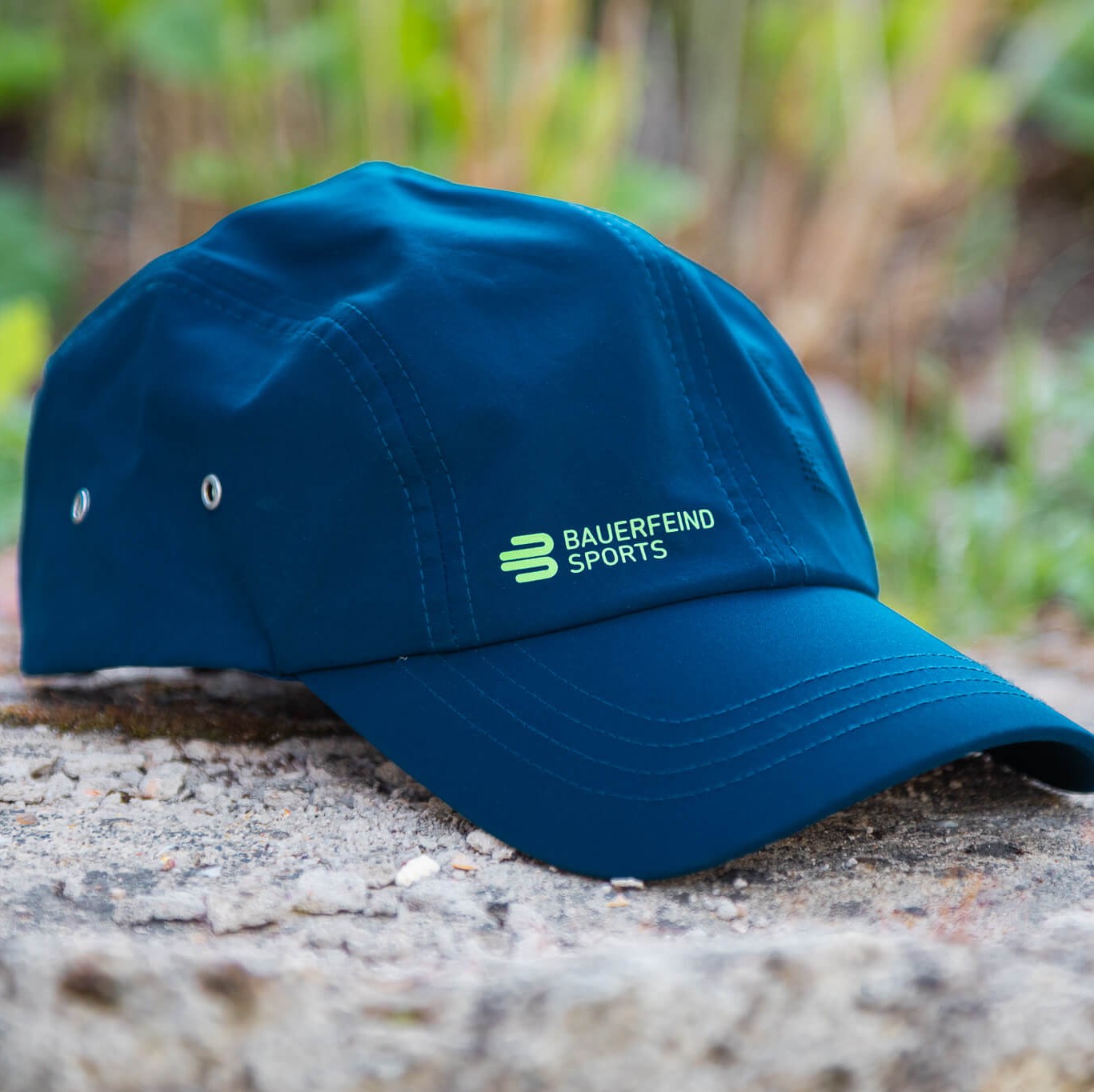 Blauwe Bauerfeind Sports pet met groen logo ligt op een steen in de buitenlucht, omgeven door onscherp groen op de achtergrond – een sportieve metgezel voor actieve dagen buiten.