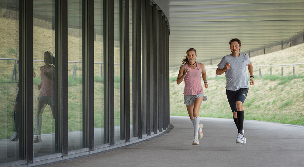 Samen sterk – kilometer na kilometer. Twee lopers – een vrouw en een man – rennen samen onder een overdekte passage. De vrouw draagt witte compressiesleeves, de man zwarte. Beiden bewegen synchroon en zien er gefocust en energiek uit.
